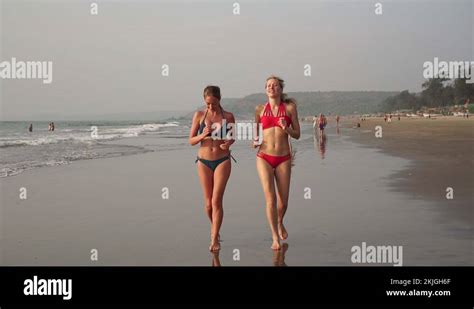 Two Beautiful Women In Bikini Swimsuits Run Barefoot Along The Seashore