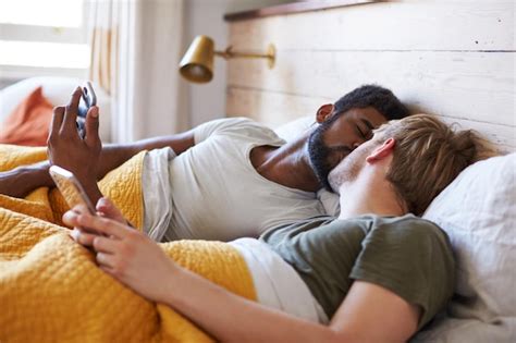 Una pareja gay masculina acostada en la cama en casa revisando teléfonos móviles juntos Foto
