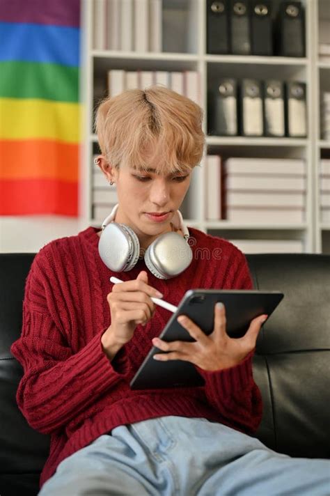 An Asian Gay Man Focuses On Something On His Tablet Screen While Sitting On The Sofa Stock Photo