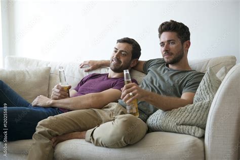Gay Male Couple On Sofa At Home Watching TV Drinking Beer Stock Photo