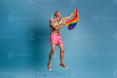 Full Length Of Mature Gay Man Carrying Rainbow Flag And Smiling While Jumping Against Blue