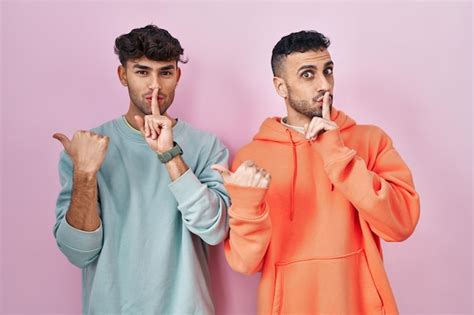 Jovem casal gay hispânico em pé sobre fundo rosa pedindo para ficar quieto o dedo nos lábios