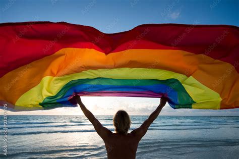Silhouette Of Man Holding Gay Pride Rainbow Flag In Front Of Tropical Beach Sunrise Scene Stock