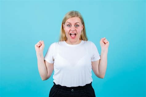 Une Fille Blonde Heureuse Regarde La Caméra Et Lève Les Poings Sur Fond Bleu Photo Gratuite