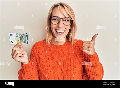 Schöne blonde Frau hält Euro Banknote lächelnd glücklich und positiv Daumen nach oben tun