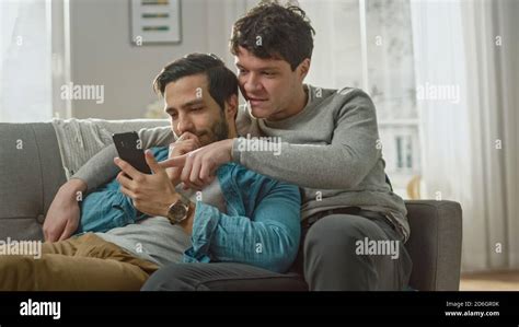 Cute Male Gay Couple Spend Time At Home They Are Lying Down On A Sofa And Use A Smartphone