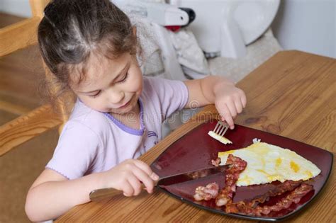 Young Pretty Girl Eating Eggs And Bacon With Knife And Fork For The