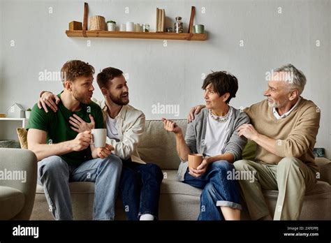 A Gay Couple Sits On A Couch With Parents Enjoying A Conversation And Warm Drinks Stock Photo