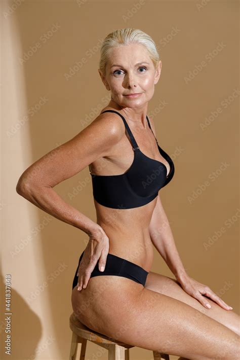 Studio Shot Of Beautiful Mature Woman In Black Underwear Looking At Camera Sitting On A Chair