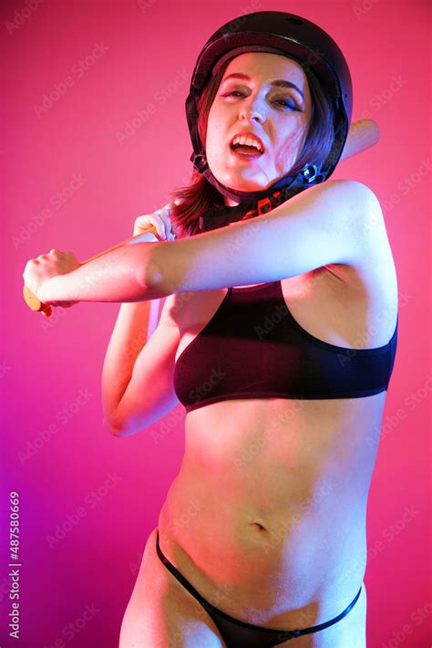Sporty Woman Posing In Bikini With Baseball Bat And Helmet In Front Of Pink Background In Studio