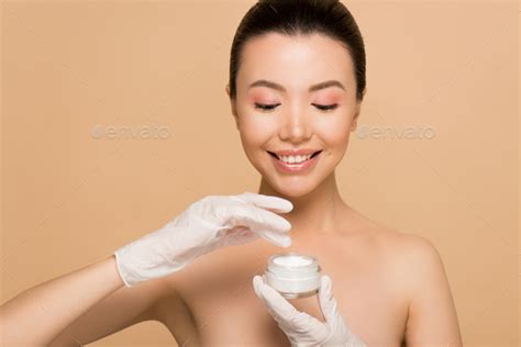 Attractive Happy Nude Asian Girl In Latex Gloves Applying Face Cream Isolated On Beige Stock
