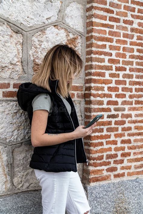 Vertical Shot Of A Blonde Spanish Girl Standing By A Brick Wall Looking At Her Phone Stock Photo