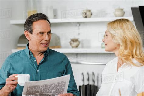 Mature Wife Looking At Husband In Kitchen He Holding Cup Of Tea And Newspaper Royalty Free