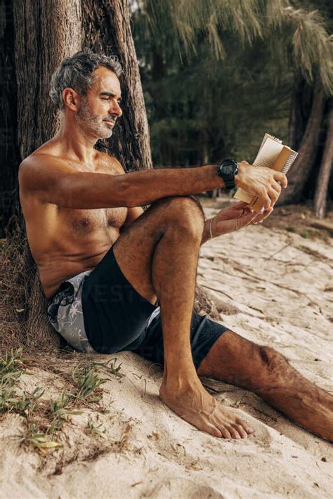 Shirtless Mature Man Reading Book While Sitting Near Tree Trunk At Beach Stock Photo
