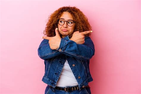 Jovem latina curvilínea isolada na parede rosa aponta lateralmente está tentando escolher entre