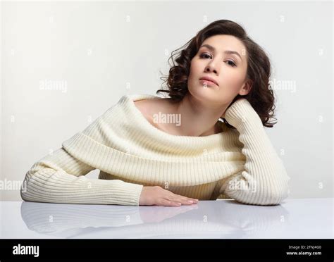 Beautiful Brunette Woman Dressed In Large White Woolen Sweater Is Sitting At White Table On Gray
