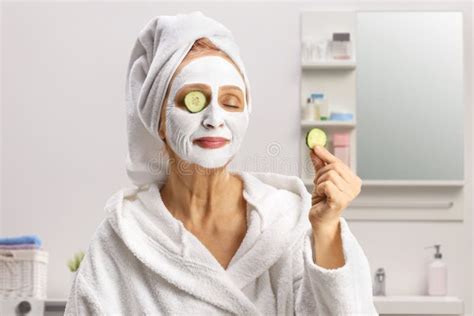 Mature Woman With A Towel On Her Head And A Face Mask Putting Cucumber On Eyes In A Bathroom