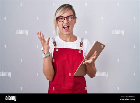 Junge blonde Frau in Kellneruniform hält Zwischenablage verrückt und verrückt Schreien und