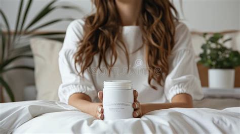 A Brunette Woman Holds A White Plastic Jar Sitting On Her Bed Showcasing A Product Stock