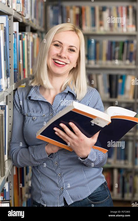 Pretty Woman With Blonde Hair Standing In The Library Blurred Books At The Back Stock Photo