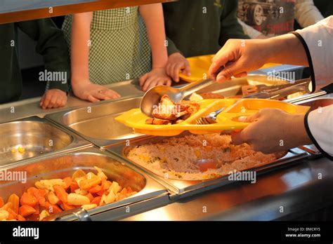 A Dinner Lady Serves Up School Dinners In A Canteen Over A Hot Plate With Pupils Waiting For