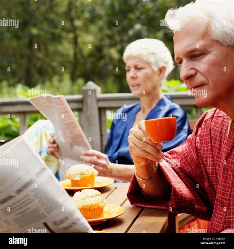 Close Up Of A Mature Man Holding A Cup And Reading A Newspaper With A Senior Woman In The