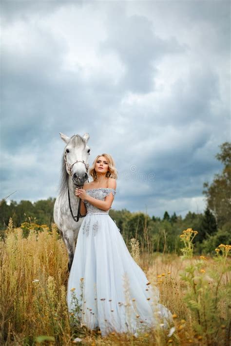 La Belle Fille Blonde Dans La Robe Frotte Un Cheval Gris Sur La Nature Dedans Photo Stock