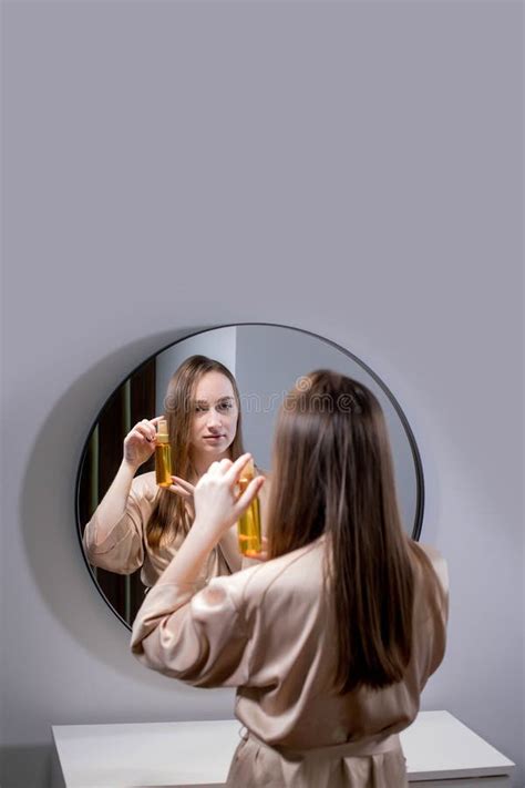 Yong Woman Brunette Applying Essential Oil Spray On Her Hair Standing Near Mirror In Bathroom