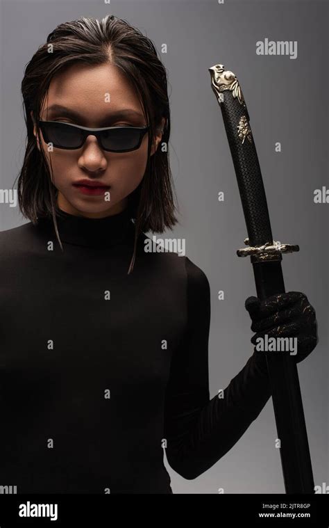 Brunette Asian Woman In Black Clothes And Stylish Sunglasses Holding Katana Sword In Scabbard