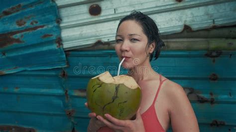 A Beautiful Girl With Wet Hair In Nude Pink Swimsuit Drinking Coconut Against A Rusty Blue Wall