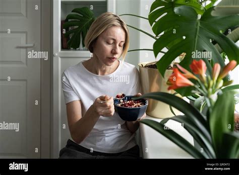 La femme blonde mature mange le petit déjeuner dans un bol assis près du seuil de la fenêtre