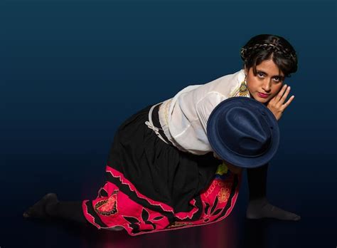 Jovem latina de vestido tradicional ajoelhado enquanto segura um chapéu azul em uma das mãos