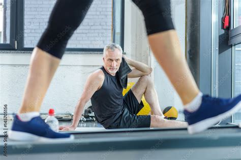 Bearded Mature Sportsman Sitting On Yoga Mat With Towel While Woman Training On Treadmill Stock