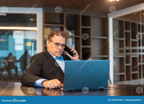 Portrait Of Mature Businessman Using Laptop Computer And Mobile Phone In Office Stock Photo
