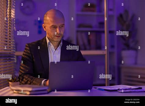 Serious Mature Man Sitting At Office Desk Working On Business Plan Using Laptop Late In Evening
