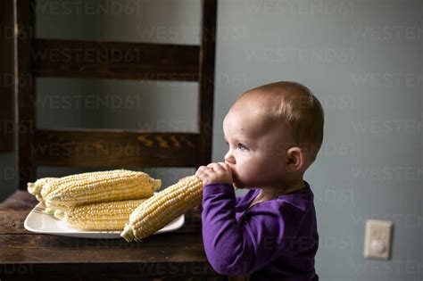 Side View Of Cute Baby Girl Eating Raw Corns At Home Stock Photo