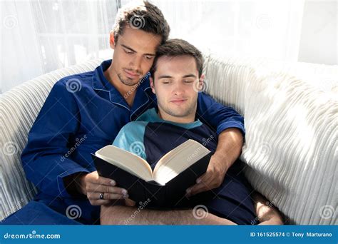Male Gay Couple Cuddling And Reading Together On Large Couch Next To