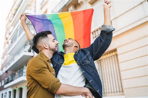 Retrato de um jovem casal gay abraçando e mostrando seu amor a bandeira do arco íris na rua