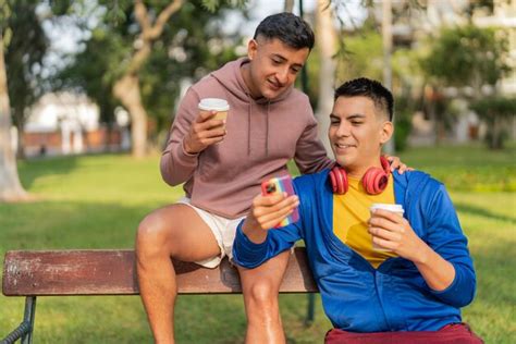 Casal gay sentado em um banco de parque usando o celular e tomando café Foto Premium