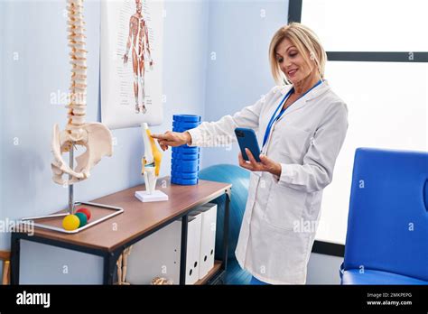 Middle Age Blonde Woman Wearing Physiotherapist Uniform Using Smartphone Pointing To Anatomical