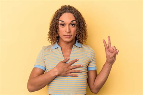 Jovem transexual latina isolada em fundo amarelo fazendo um juramento colocando a mão no peito