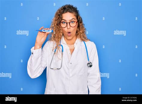 Junge schöne blonde Ärztin mit blauen Augen tragen Stethoskop halten Thermometer erschrocken in