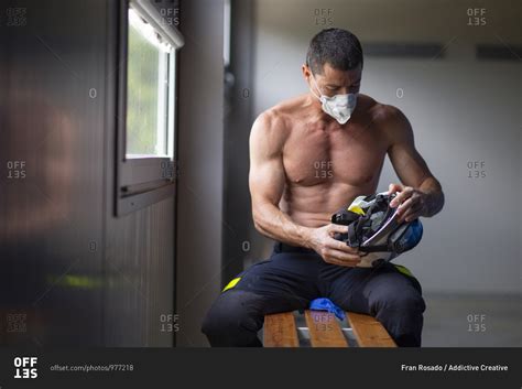 Strong Mature Male Firefighter With Naked Torso Sitting On Bench In Mask And Holding A Helmet