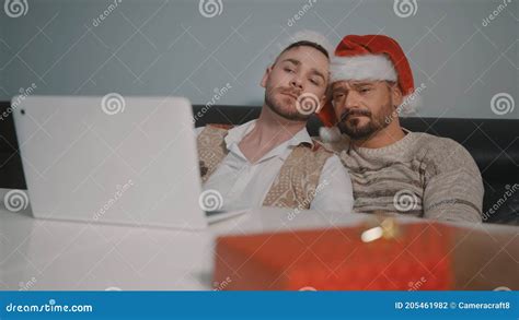 Happy Gay Male Couple With Santa Claus Hats Cuddling And Watching Movie On The Christmas Eve
