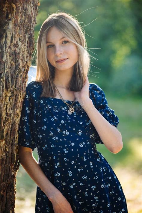 Beautiful Blonde Teenager Posing In Summery Dress Leaning Casually Against Wooden Tree Trunk