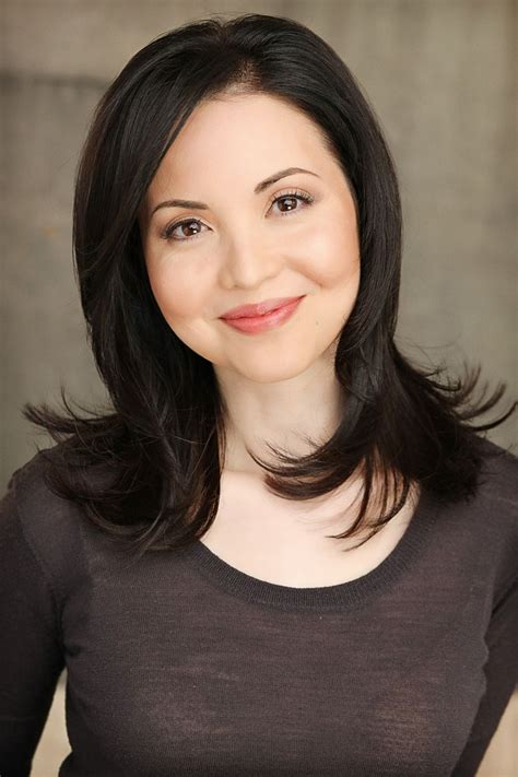 A Woman With Dark Hair And Brown Shirt Smiling In Front Of A Gray Wall