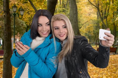 Two Girlfriends Walk In The Autumn Park And Take Selfie In Phone Stock