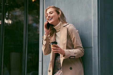 Belle fille blonde souriante en trench coat élégant avec du café pour parler joyeusement sur un