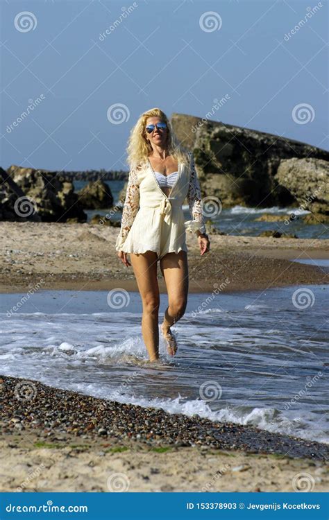 Tanned Blonde In White Dress And Sunglasses Walks On The Water Of The Baltic Sea On A Sunny Day