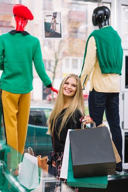 Souriante blonde jeune femme debout devant une vitrine tenant des sacs shopping colorés Photo